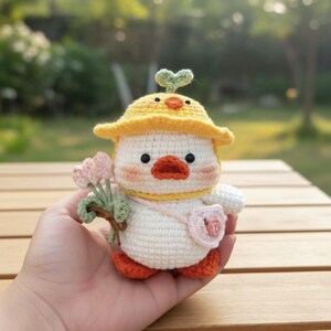 May include: A handmade crochet duckling doll. It has a yellow hat, orange beak and feet, and a small pink purse. The duckling is holding a bouquet of pink flowers. The doll is white with peach-colored cheeks.