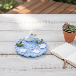 May include: A handmade crochet coaster featuring a white duck with a yellow beak, sitting on a blue pond with white flowers and green accents. The coaster is placed on a white wooden surface, with a succulent in a terracotta pot in the background.