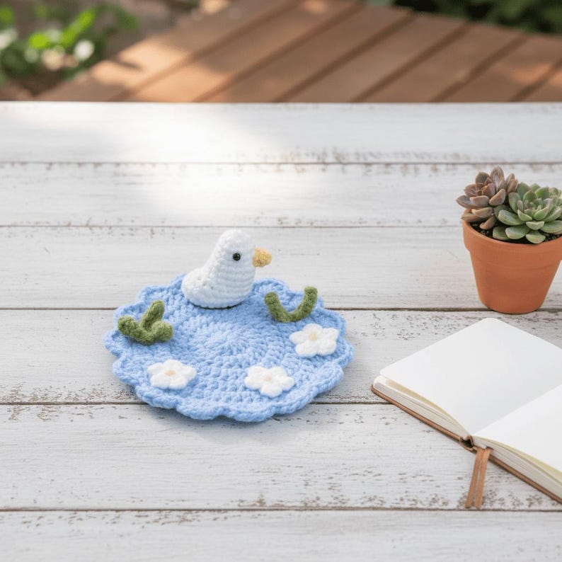 May include: A handmade crochet coaster featuring a white duck with a yellow beak, sitting on a blue pond with white flowers and green accents. The coaster is placed on a white wooden surface, with a succulent in a terracotta pot in the background.