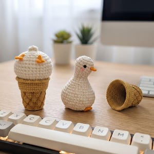 May include: A white crocheted duck figurine, a white crocheted ice cream scoop in a tan waffle cone, and an empty tan crocheted waffle cone. The duck has small orange feet, a small orange beak, and pink blush marks on its cheeks.