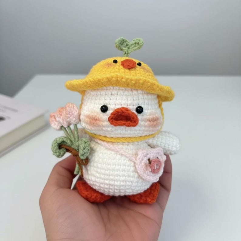 May include: A crocheted duck toy, white with orange feet and beak, wearing a yellow hat with a green sprout. It holds pink flowers and a pink purse. The duck has black eyes and peach-coloured cheeks.