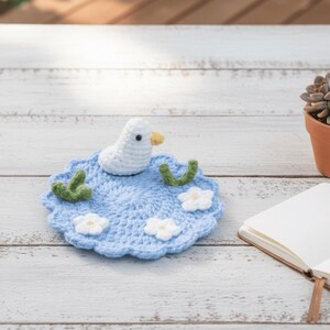 May include: A handmade crochet coaster featuring a white duck on a blue pond with white flowers and green accents. The coaster is round and sits on a white wooden surface. A small succulent in a terracotta pot is in the background.