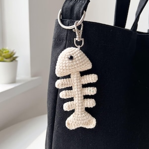 May include: A cream-colored crocheted fish skeleton keychain attached to a black canvas tote bag with a silver-toned metal lobster clasp and a circular ring. The fish skeleton features a single black bead eye and a textured yarn construction.