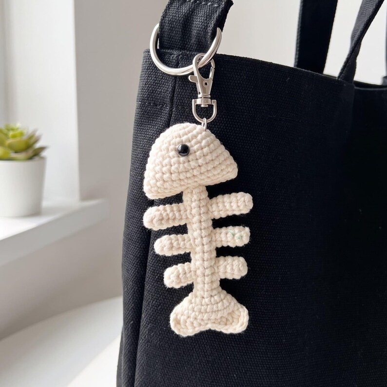 May include: A cream-colored crocheted fish skeleton keychain attached to a black canvas tote bag with a silver-toned metal lobster clasp and a circular ring. The fish skeleton features a single black bead eye and a textured yarn construction.