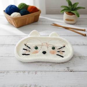 May include: A white, crocheted cat-shaped tray holding a pair of eyeglasses. The tray has a cat face design with black whiskers, a black nose, and pink cheeks. A basket of yarn and a small crocheted plant are in the background.