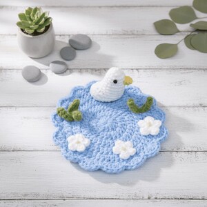 May include: A handmade crochet coaster featuring a white duck on a light blue pond with white flowers and green accents. The coaster is round with a scalloped edge, perfect for a cup or glass. The coaster measures approximately 5 inches in diameter.