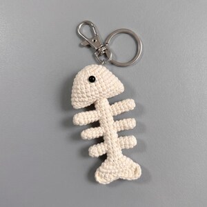 May include: A cream-colored crocheted fish skeleton keychain featuring a single black bead for an eye. The fish bone design is attached to a silver-toned metal lobster clasp and a circular split ring, providing a secure way to attach the item.