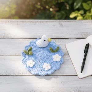 May include: A handmade, light blue crochet coaster with a white duck, green leaves, and white flowers. The duck has a yellow beak. The coaster is on a white wooden surface, next to a notebook and pen.