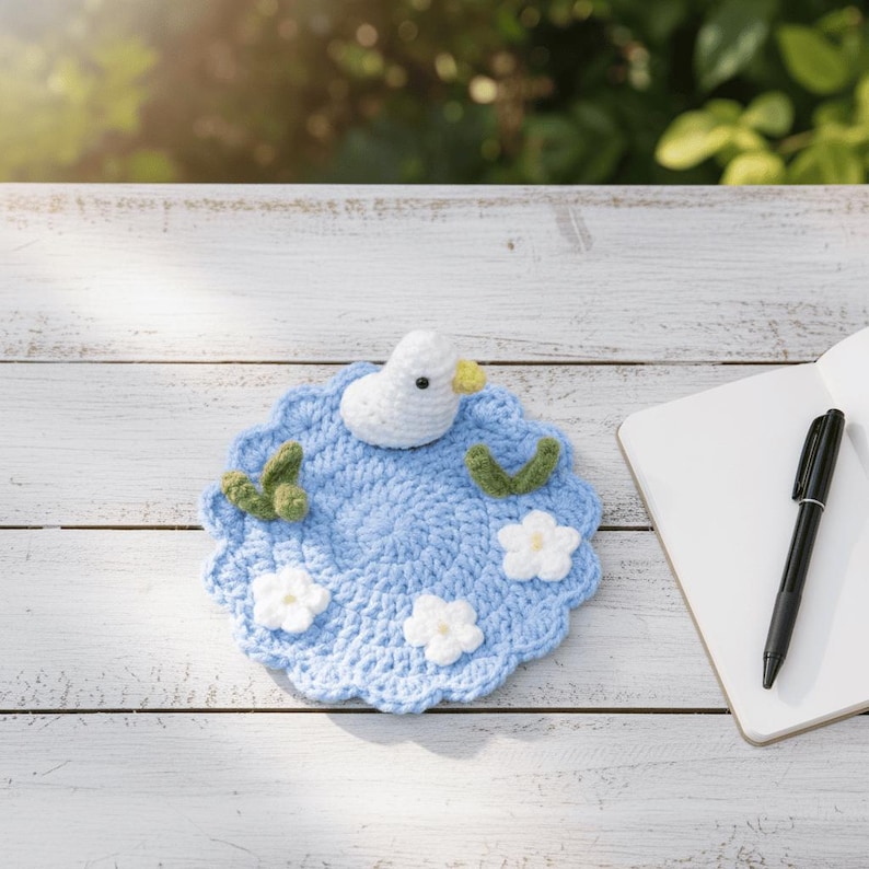 May include: A handmade, light blue crochet coaster with a white duck, green leaves, and white flowers. The duck has a yellow beak. The coaster is on a white wooden surface, next to a notebook and pen.