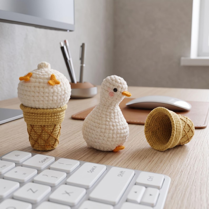 May include: A crocheted white duck with a small orange beak and pink cheeks, positioned next to a crocheted white ice cream scoop with orange accents inside a yellow waffle cone, and an empty yellow waffle cone lying on its side.