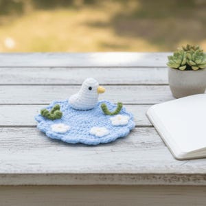 May include: A handmade crochet duckling on a light blue lily pad with white flowers and green accents. The duckling is white with a yellow beak. The lily pad is on a white wooden surface, with a succulent plant and a notebook in the background.