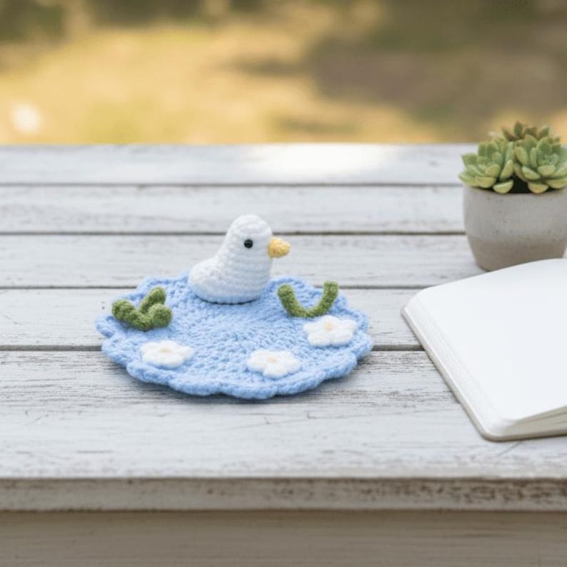 May include: A handmade crochet duckling on a light blue lily pad with white flowers and green accents. The duckling is white with a yellow beak. The lily pad is on a white wooden surface, with a succulent plant and a notebook in the background.
