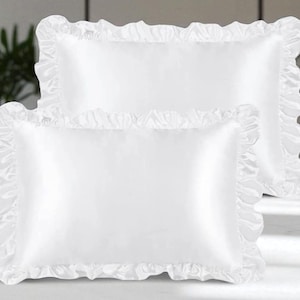 May include: Two white rectangular pillowcases with frilled edges. The pillowcases are made of a smooth, satin-like material. The pillows are displayed on a light-coloured surface, with plants in the background.