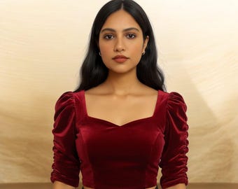 May include: A burgundy velvet crop top with a square neckline and three-quarter sleeves. The sleeves are gathered at the elbow, adding volume. The top is a solid color and has a fitted silhouette.