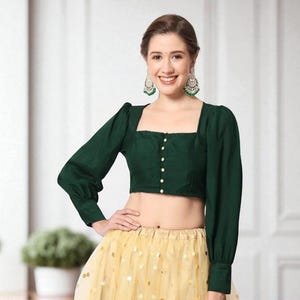 May include: A dark green crop top with long sleeves and a square neckline. The top has a row of small white buttons down the front. The model is wearing a yellow skirt with gold sequins and green earrings.
