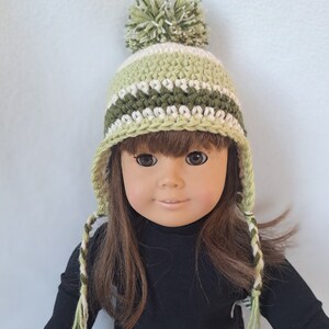 May include: A green and white striped crocheted hat with a white pom pom on top. The hat has long ties that hang down the sides. The hat is being worn by a doll with brown hair.