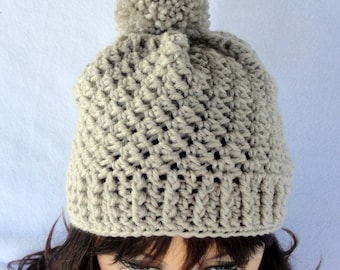Op de afbeelding: Een lichtbeige gehaakte beanie-muts met een gestructureerd steekpatroon en een bijpassende pompon bovenop. De muts heeft een geribbelde boord. Deze handgemaakte muts is ontworpen voor warmte en stijl.