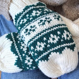 May include: A pair of white knitted mittens with a green geometric pattern. The mittens are folded and placed on a blue denim surface.