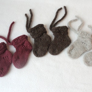 Pode incluir: Seis pares de botinhas de bebé de malha em tons de bordô, castanho e cinza. Cada par tem uma bainha dobrada e laços presos. As botinhas estão dispostas sobre uma superfície branca, mostrando o artesanato feito à mão.