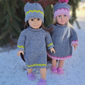 May include: Two dolls dressed in matching gray knit dresses and hats. The dresses have yellow zigzag trim on the sleeves and hem. One doll wears a gray hat with yellow zigzag trim, and the other wears a gray hat with pink zigzag trim and a pink pom-pom. Both dolls wear purple clogs.
