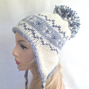 Puede incluir: Un gorro de punto blanco y azul con un pompón en la parte superior y orejeras. El gorro tiene un patrón de rayas y está hecho de un hilo suave y cálido.