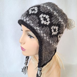 Puede incluir: Un gorro de punto negro y blanco con un patrón geométrico y orejeras. El gorro tiene un pompón en la parte superior y borlas en las orejeras.