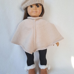 Puede incluir: Una muñeca con un gorro de punto beige, un poncho de punto beige, leggings negros y botas marrones con forro de piel sintética.