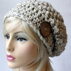 Op de afbeelding: Een witte gebreide beanie met een bruine knoop. De beanie heeft een slouchy pasvorm en is gemaakt van een getextureerd garen.