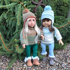 Pode incluir: Duas bonecas vestindo roupas de inverno. Uma boneca está vestindo um suéter verde, calças verdes e um chapéu de tricô marrom com uma pompom. A outra boneca está vestindo um suéter branco com um padrão azul e branco, calças azuis e um chapéu de tricô azul com uma pompom.