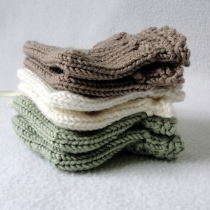 May include: A stack of four knitted items in varying neutral colors. The stack includes items in shades of beige, cream, and green. The items appear to be small, possibly baby booties or similar accessories. The texture of the knitwear is visible.