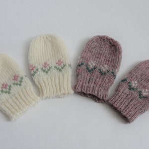 May include: Four knitted baby mittens, two white with pink and green floral patterns and two pink with white and green floral patterns.