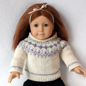May include: A doll wearing a white knitted jumper with a purple and grey geometric pattern. The jumper has a high neck and long sleeves.
