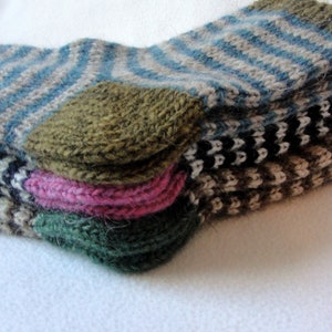 K&ouml;nnte beinhalten: Ein Stapel von vier gestrickten Socken in verschiedenen Farben und Mustern, darunter blau-wei&szlig;e Streifen, schwarz-wei&szlig;e Streifen, braun-wei&szlig;e Streifen und eine einfarbige gr&uuml;ne Socke.