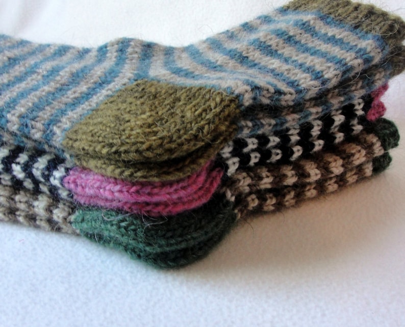 K&ouml;nnte beinhalten: Ein Stapel von vier gestrickten Socken in verschiedenen Farben und Mustern, darunter blau-wei&szlig;e Streifen, schwarz-wei&szlig;e Streifen, braun-wei&szlig;e Streifen und eine einfarbige gr&uuml;ne Socke.