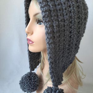 Peut inclure: Une cagoule grise au crochet avec deux pompons. La cagoule est faite d'un fil épais et texturé et présente un sommet pointu. Les pompons sont attachés aux extrémités des rabats de la cagoule. La cagoule est présentée sur une tête de mannequin.