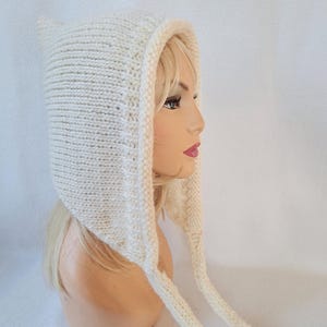 Puede incluir: Gorro tejido de color crema con orejas puntiagudas y lazos largos. La capucha está hecha de un hilo grueso y texturizado y está diseñada para cubrir la cabeza y el cuello. Un accesorio acogedor y elegante para el frío.