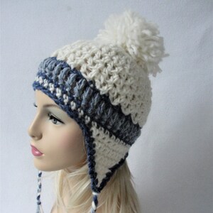 Könnte beinhalten: Weiße und blaue Häkelmütze mit Ohrenklappen und einem weißen Bommel oben.