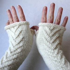 May include: A pair of ivory-colored knitted fingerless gloves. The gloves feature a textured chevron pattern and a ribbed cuff. The design is a cozy, handmade accessory, perfect for adding warmth and style. The gloves are a neutral color.