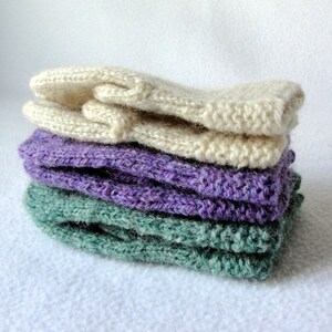 May include: A stack of four knitted fingerless gloves in different colors. The top glove is cream, the second is purple, the third is green, and the bottom glove is cream.