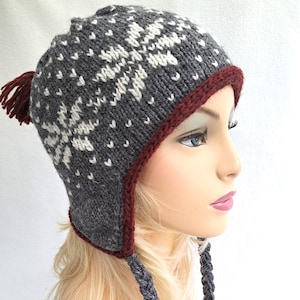 Puede incluir: Gorro de invierno de punto gris con un patrón de copo de nieve blanco y un ribete burdeos. El gorro tiene orejeras y un pompón en la parte superior.