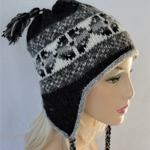 Puede incluir: Un gorro de invierno tejido en tonos negro, gris y blanco. El gorro presenta un patrón geométrico y orejeras con lazos trenzados. Una borla adorna la parte superior. El gorro está diseñado para mantener abrigado al usuario en climas fríos.