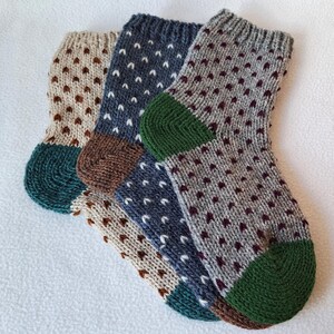 May include: Three pairs of knitted socks with different patterns. The socks are brown, grey, and blue with white accents. The socks have green heels and toes.