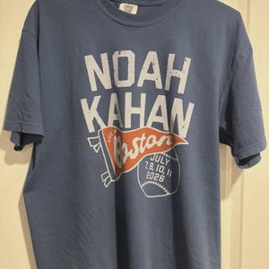 May include: A blue short-sleeved t-shirt featuring white and orange graphic text that reads NOAH KAHAN, LIVE IN Boston, and JULY 7, 8, 10, 11 2026. The design includes an orange pennant flag and a white baseball graphic on the front of the garment.