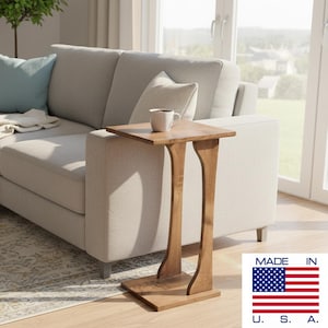 May include: A light brown wooden C-shaped side table with a square top and a curved support base. The table features a smooth finish and is positioned next to a light gray sofa. A white ceramic mug sits on the table surface. The text MADE IN U.S.A. is visible.