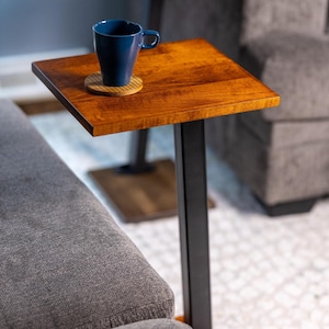 May include: A square, wooden side table with a dark stain finish. The table has a black metal frame and a dark blue mug on top, resting on a wooden coaster. The table is positioned next to a gray sofa.