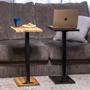 May include: Two side tables with square wooden tops and black metal legs. One table holds a blue mug and a plate with cookies, the other holds a laptop. The tables are designed to be placed next to a sofa.