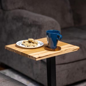 May include: A small, square wooden side table with a black metal base. On the table is a blue mug on a wooden coaster and a small plate with chocolate chip cookies. The table has a natural wood grain finish.