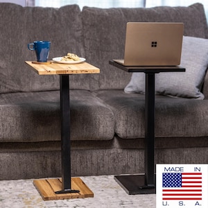 May include: Two small, square side tables with black metal legs. One table has a light wood top with a blue mug and a plate of cookies. The other table has a dark wood top with a laptop. The tables are next to a gray couch.