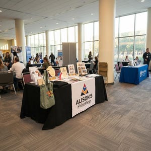 May include: A convention hall with multiple vendor tables. One table has a black tablecloth with a white banner that reads "Alison's Printshop." Various printed materials and a tote bag are displayed. Other tables are covered in blue.