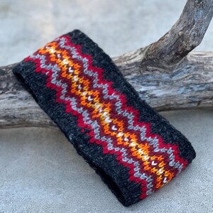 May include: A dark gray knitted headband with a zig-zag pattern in red, orange, yellow, white, and gray. The headband is made of a soft, textured material and is designed to keep the ears warm. The accessory is suitable for cold weather.